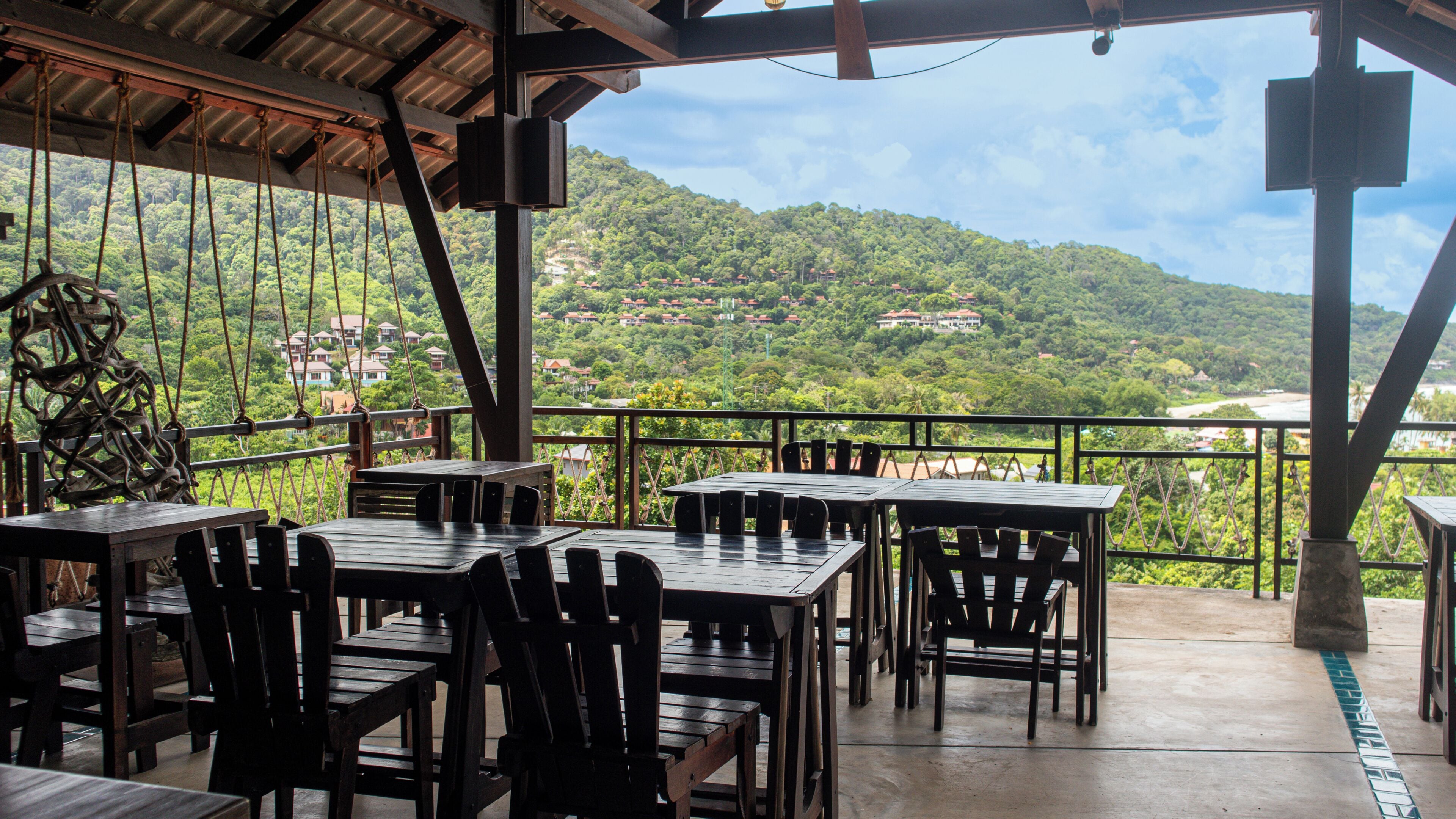Un área de comedor al aire libre con mesas y sillas, con vistas a una colina verde y exuberante con casas.