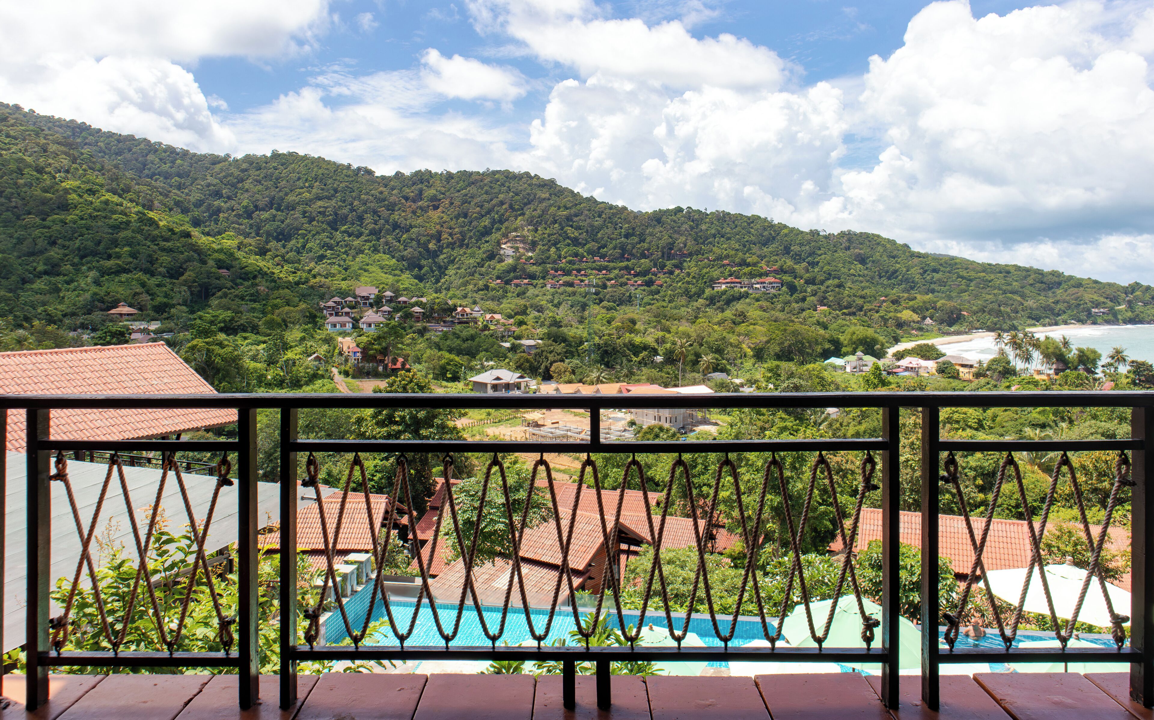 Blick auf einen Swimmingpool mit einer Holzterrasse im Vordergrund, eine gebirgige Landschaft im Hintergrund und einem klaren Himmel.