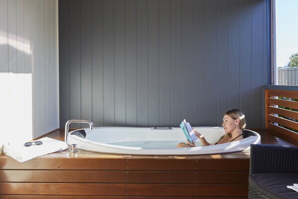A person relaxing in a hot tub with a book, wooden floor, and a window with blinds.