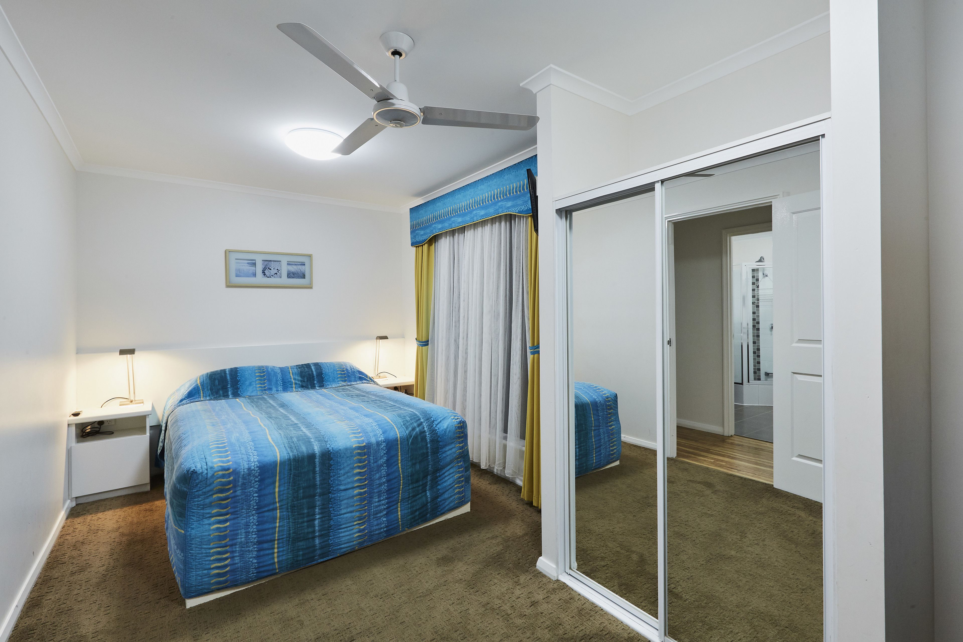 Habitación de hotel con cama, mesita de noche, lámpara, ventilador de techo y una puerta corredera que da a otra habitación.
