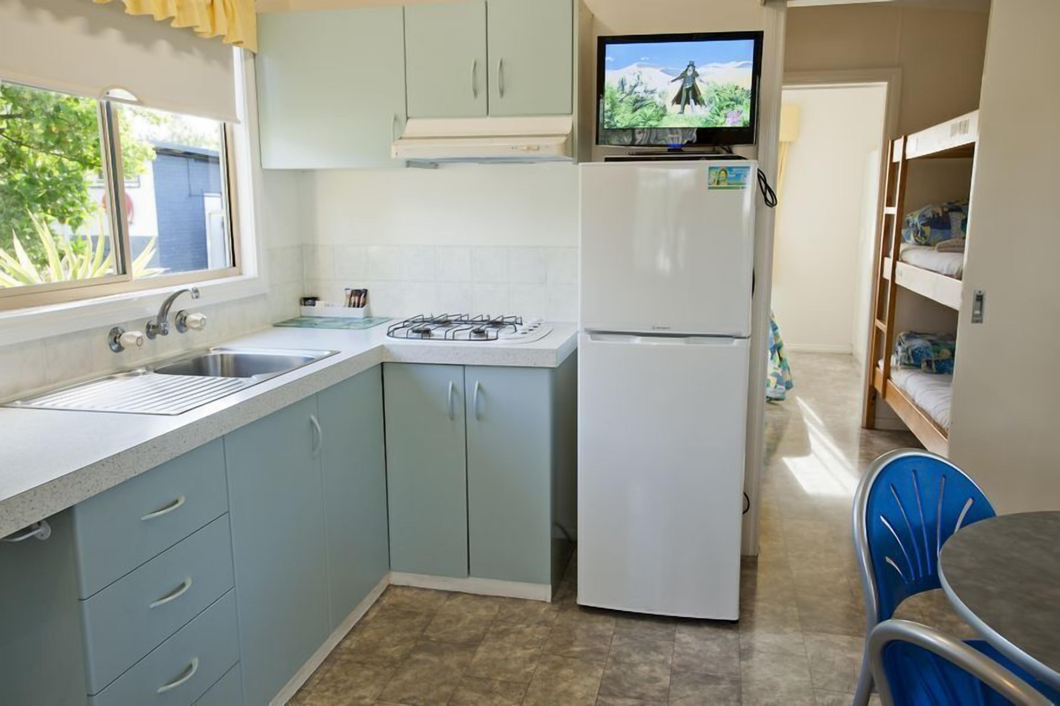 A compact kitchen with a sink, stove, and refrigerator, a small dining area with a table and chairs, and a bunk bed in the background.