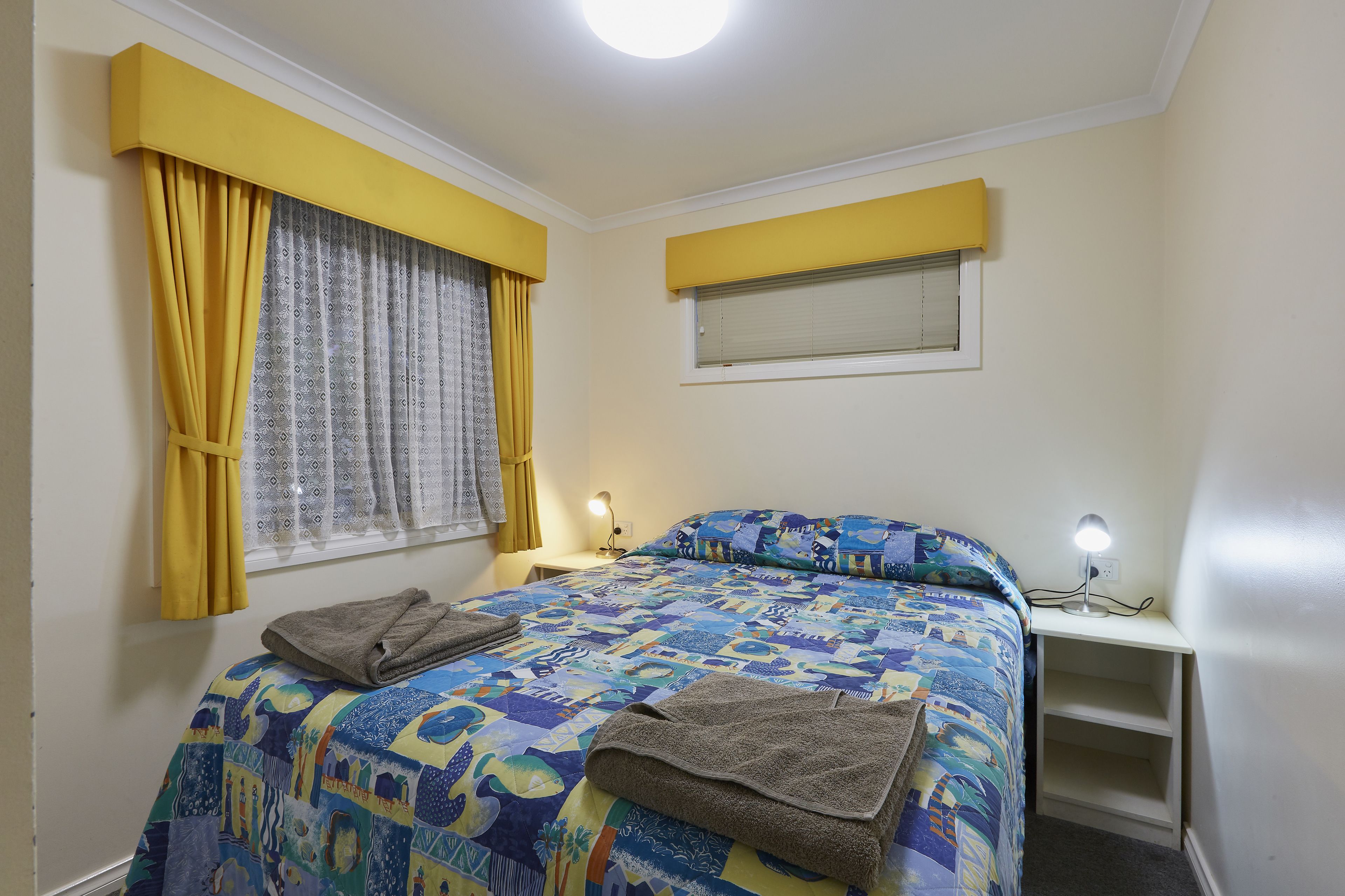 Un dormitorio con cama, mesita de noche, lámpara, ventana con cortinas y estante.
