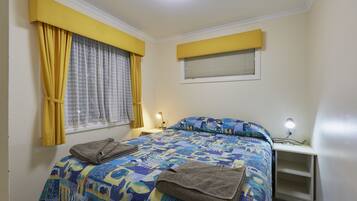 Un dormitorio con cama, mesita de noche, lámpara, ventana con cortinas y estante.