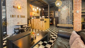 A modern interior with a checkered floor, a wooden reception desk, and a seating area with a black leather couch and a table with a vase of flowers. - Na Siam Guesthouse (Phuket)