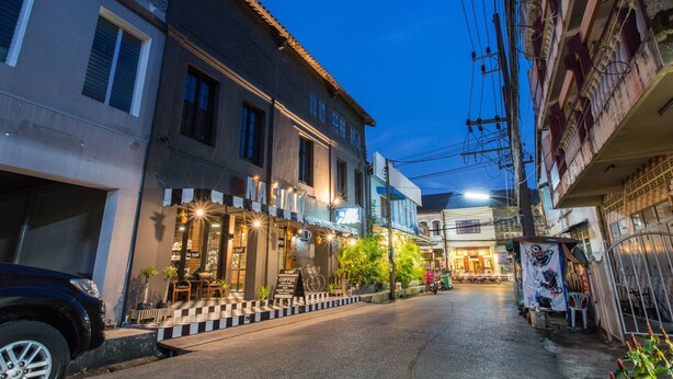 A street view at dusk with a café named "NASSIA" on the left, a parked car, and a building with a balcony on the right. - Na Siam Guesthouse (Phuket)