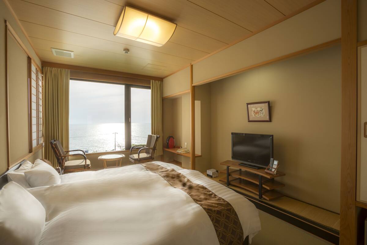Ett hotellrum med en stor säng, en tv, ett skrivbord och en stol. Det finns ett fönster med utsikt över havet.