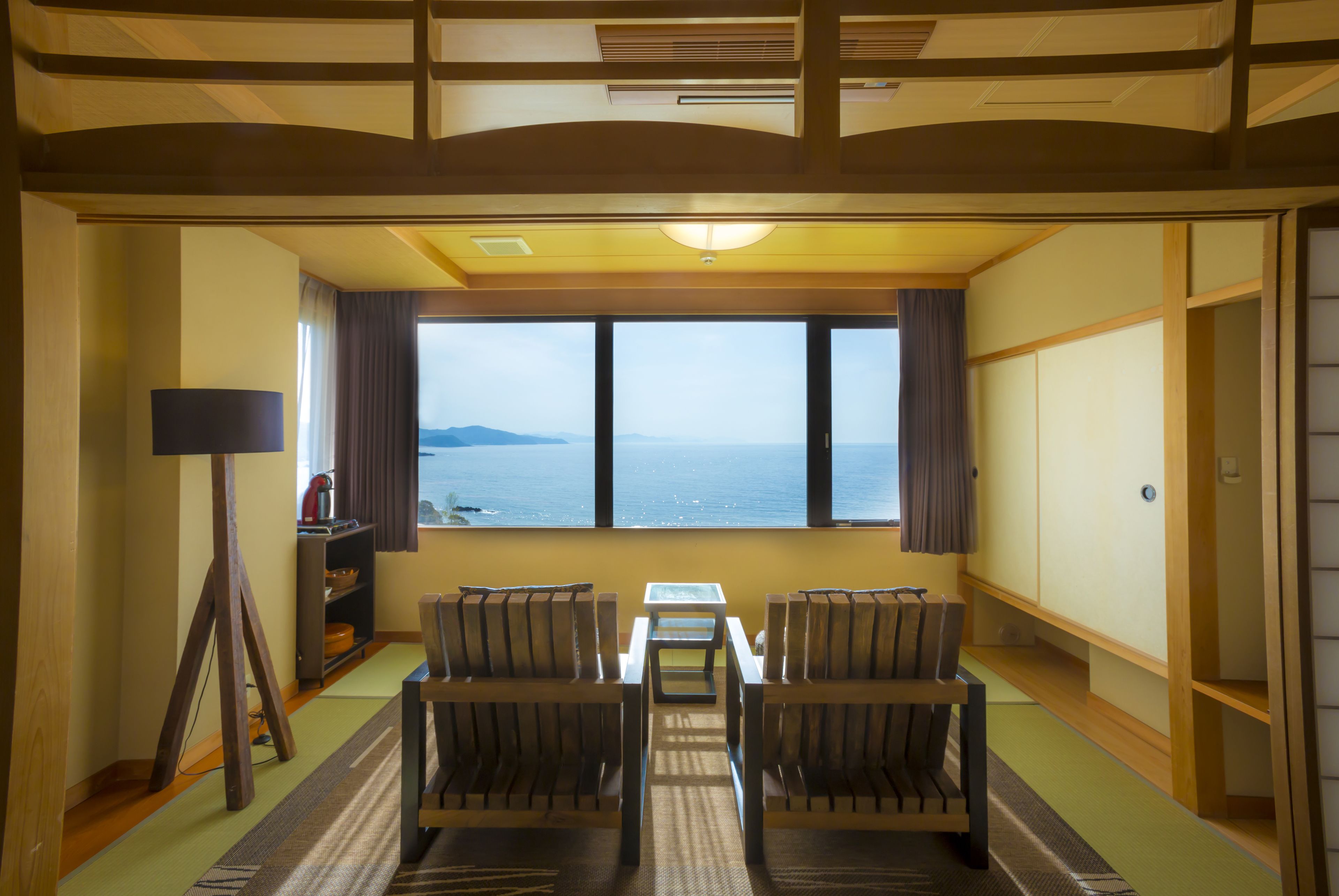 A room with a view of the sea, a small table, two chairs, a lamp, and a shelf with items.