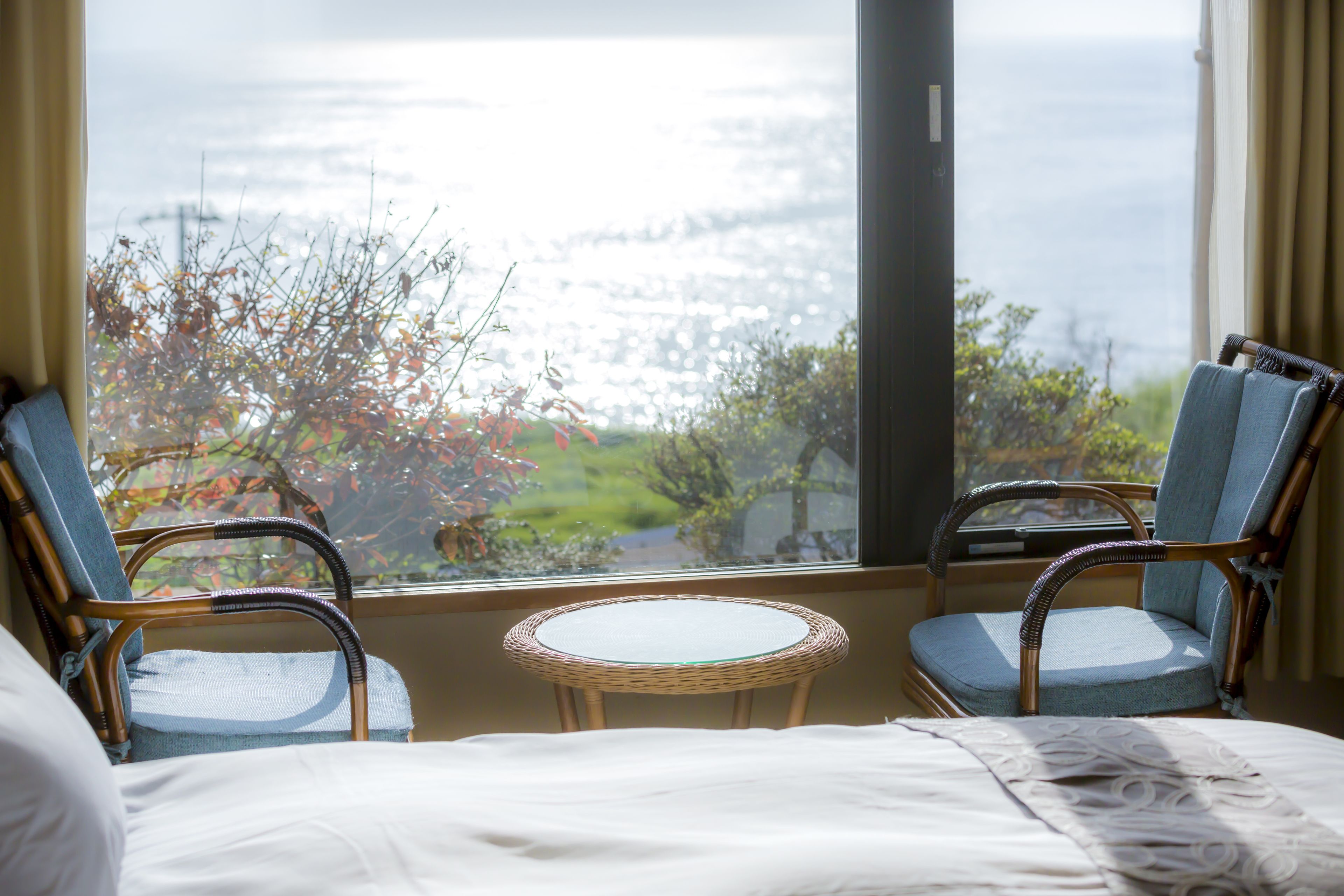 A room with a bed, two wicker chairs, a small round table, and a large window offering a view of trees and a body of water.