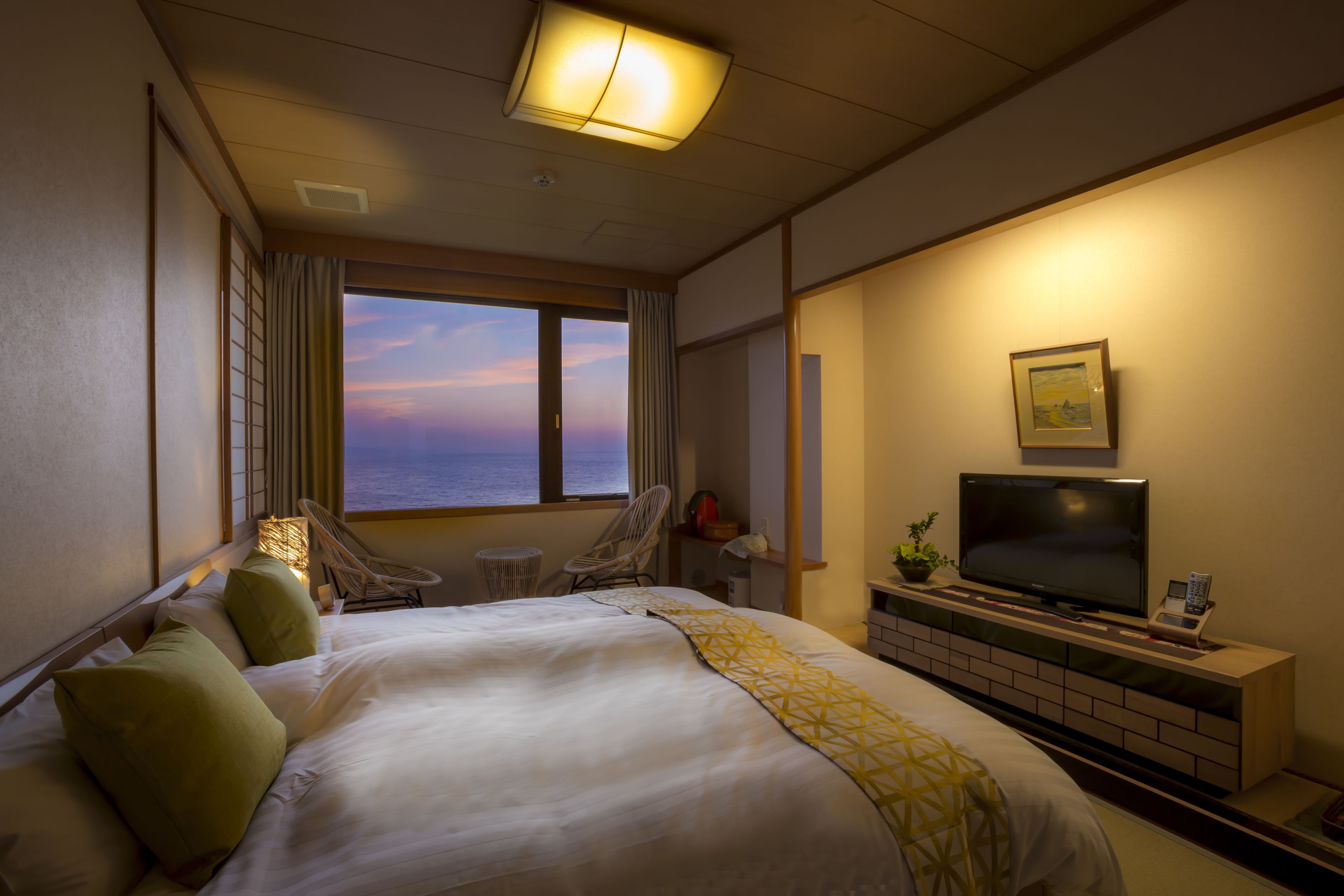 A hotel room with a bed, a TV, a desk, a chair, and a view of the sunset.