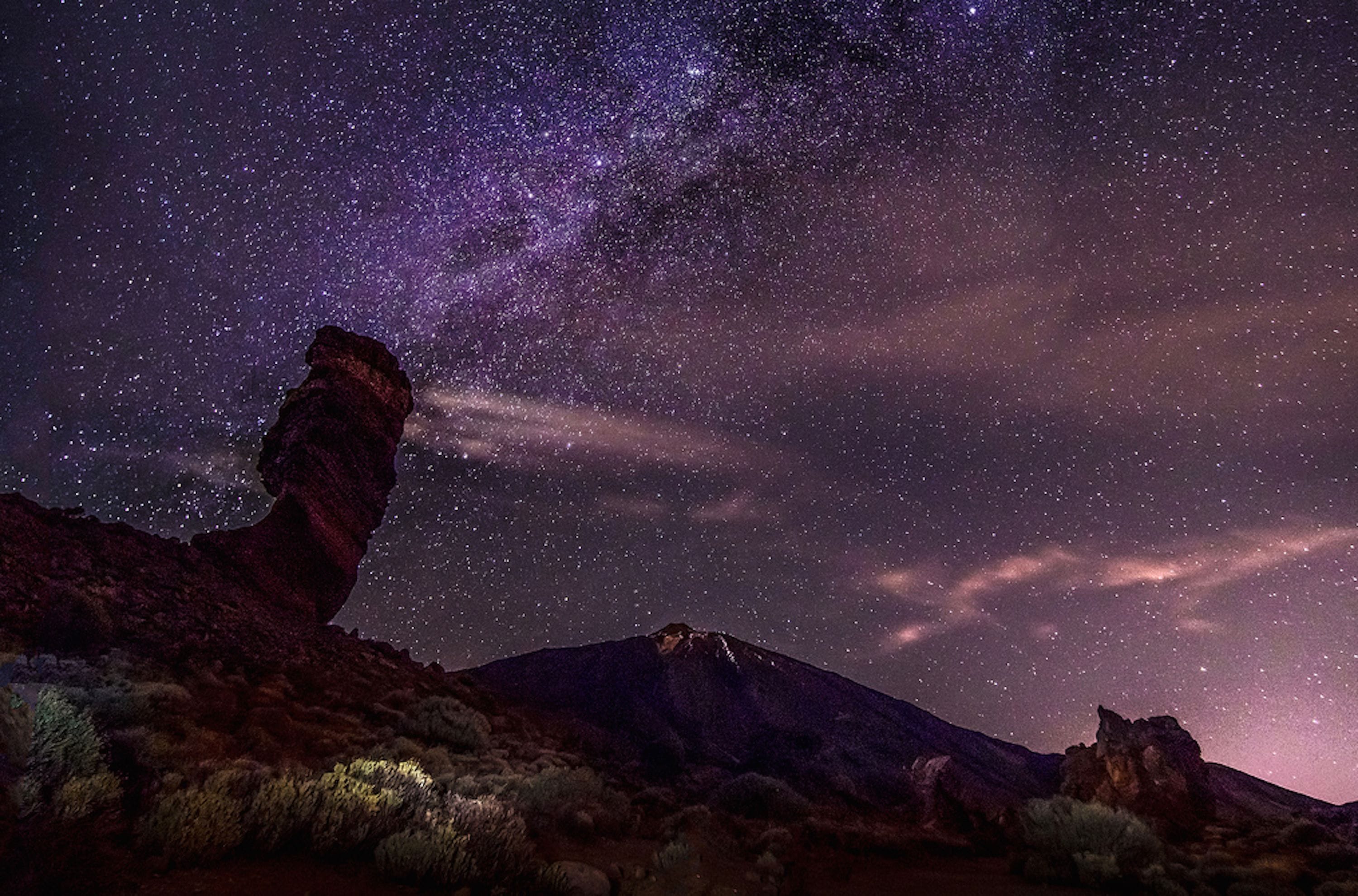 A starry night sky over a mountainous landscape with a prominent rock formation.