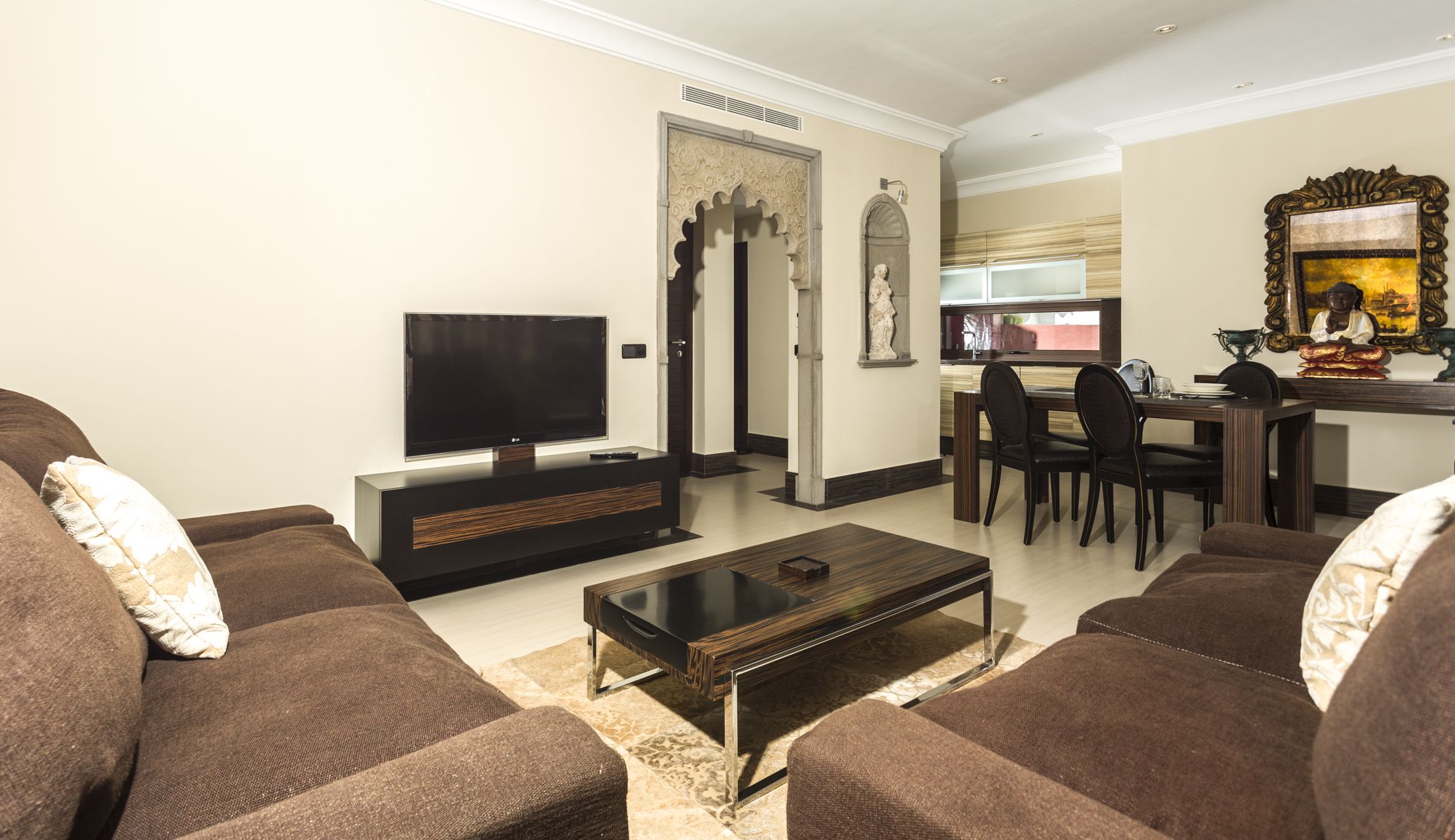 A living room with brown sofas, a flat-screen TV, a coffee table, and a dining area with chairs and a table.