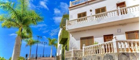 A two-story building with a white facade, a stone wall, and a balcony with a balustrade.