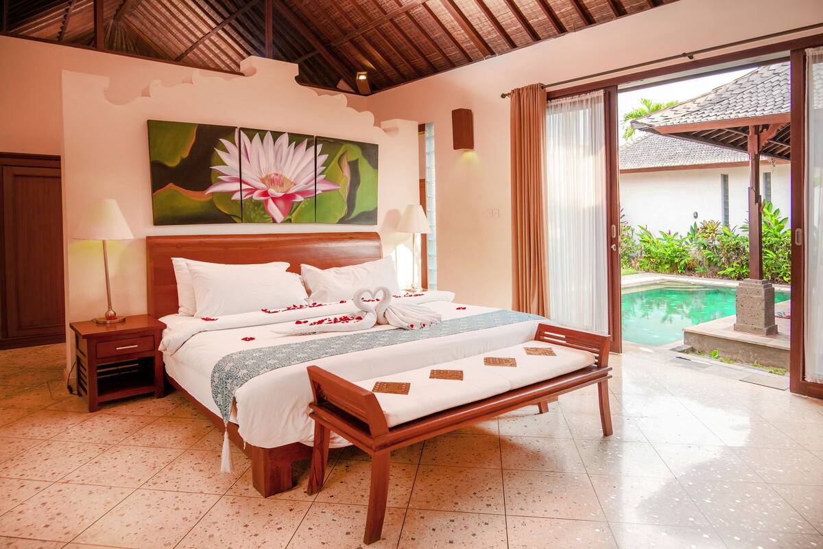 A well-lit bedroom with a large bed, a bench, and a view of an outdoor swimming pool.