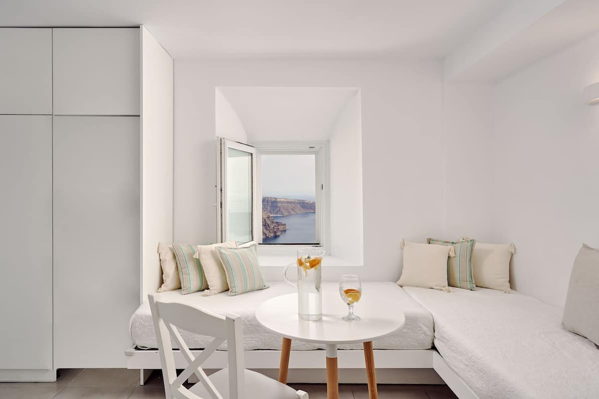 Une pièce moderne et minimaliste, avec un canapé blanc, une petite table ronde et une vue sur la mer par une fenêtre ouverte.