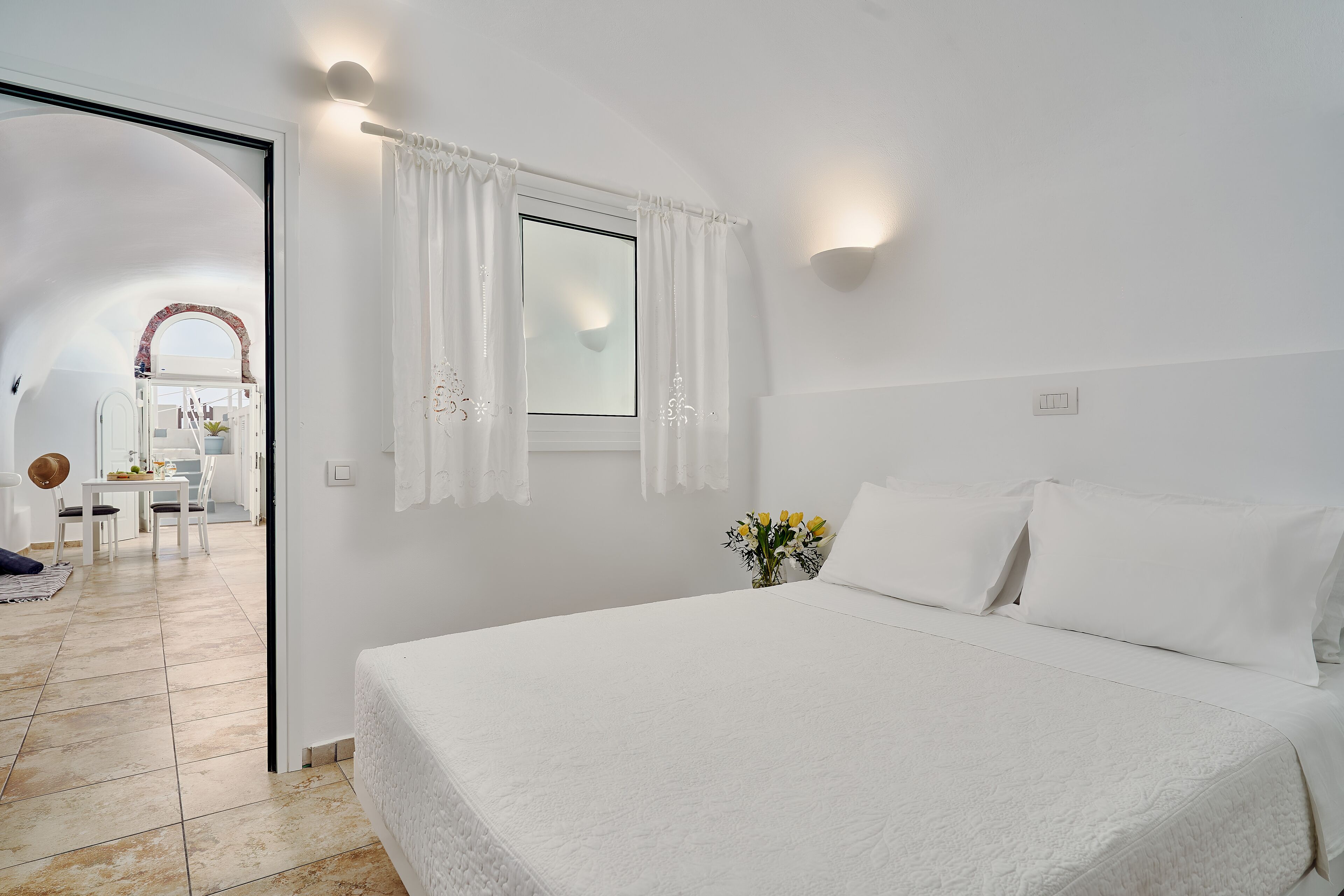 A bedroom with a large bed, white curtains, a window, and a view of a dining area.