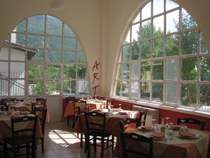 Un comedor con mesas dispuestas para comidas, amplias ventanas en arco y vistas al exterior con vegetación.