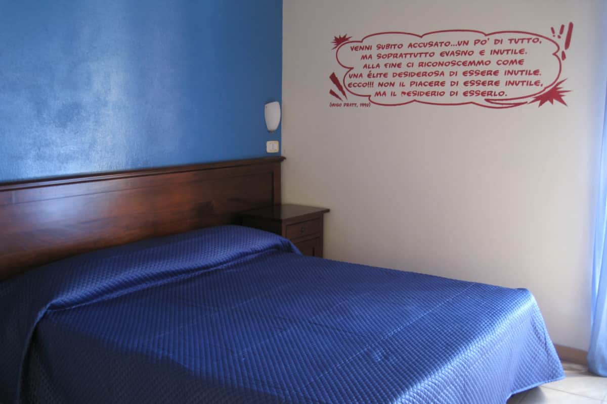 A bedroom with a wooden headboard, a single bed with a blue bedspread, a small bedside table, and a wall with a motivational quote in Italian.