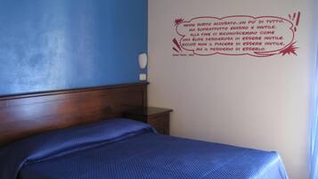 Une chambre à coucher avec une tête de lit en bois, un lit simple recouvert d’une couverture bleue, une petite table de chevet et un mur orné d’une citation motivante en italien.