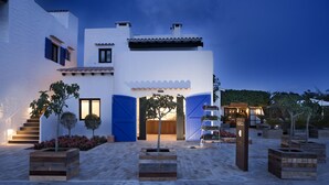 A modern building with a white facade, blue doors, and a paved courtyard with wooden planters and a staircase. - Paraíso de los Pinos Apartamentos (Formentera)