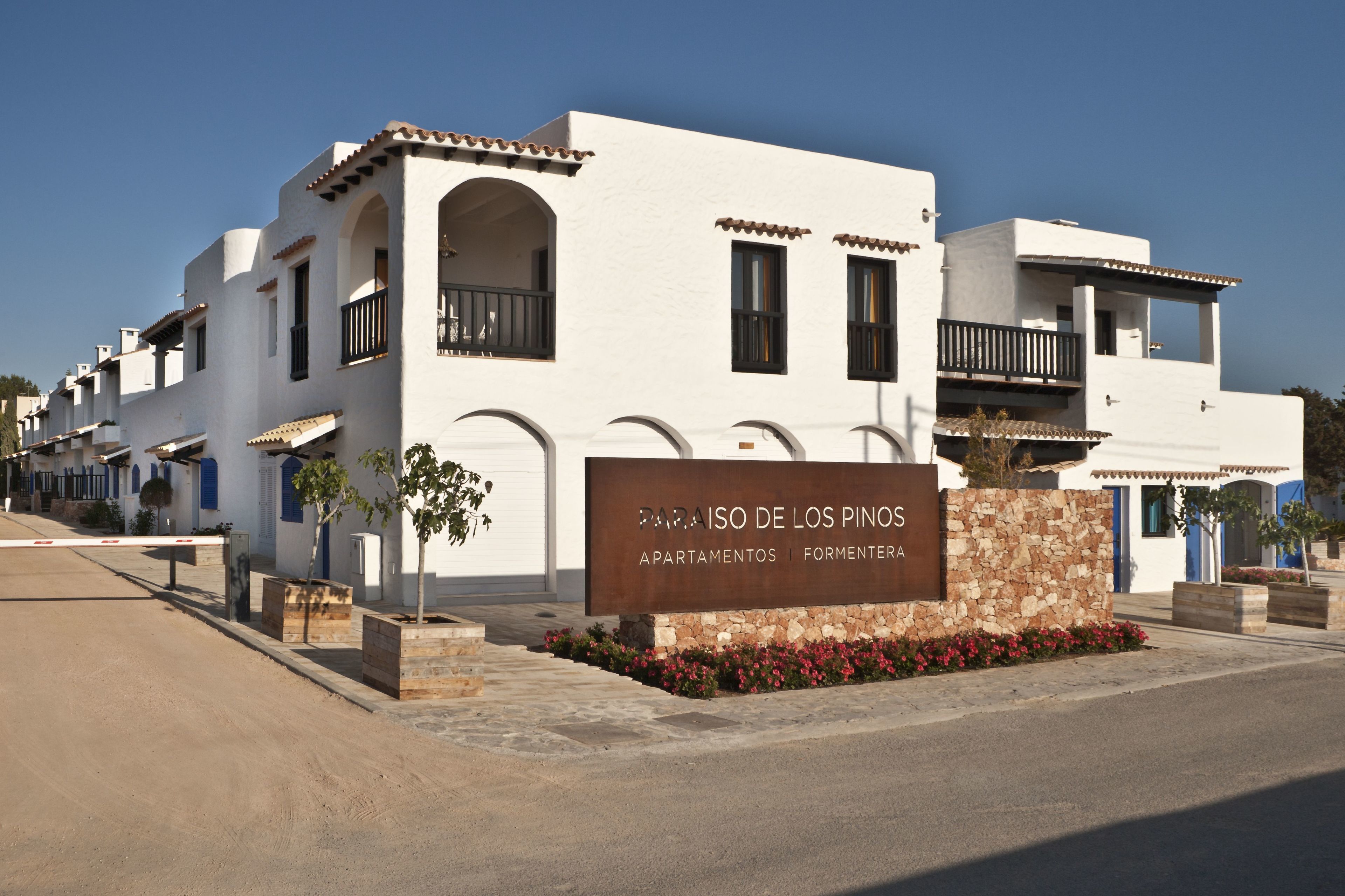 Eine Reihe weißer Gebäude mit Balkonen, ein Schild mit der Aufschrift "PARAISO DE LOS PINOS APARTAMENTOS | FORMERTEA" und eine Steinmauer mit roten Blüten.