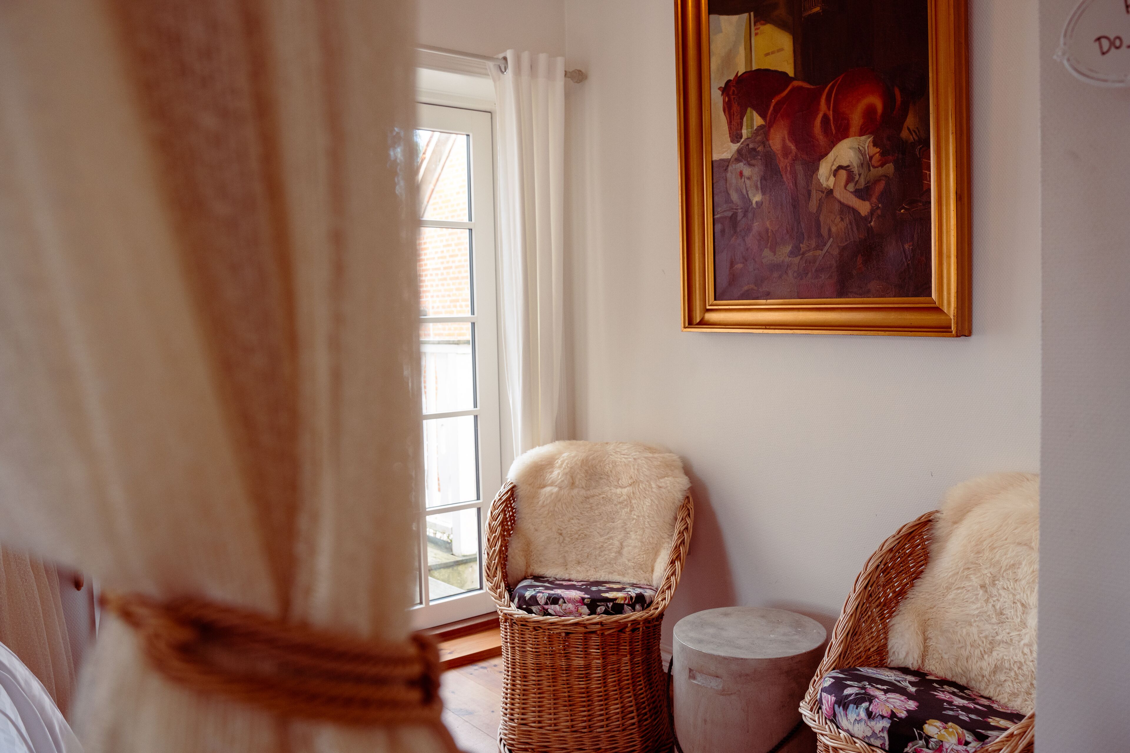A room with two wicker chairs, one with a floral cushion and another with a fur throw. A painting hangs on the wall, and a window with curtains is visible.
