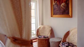 A room with two wicker chairs, one with a floral cushion and another with a fur throw. A painting hangs on the wall, and a window with curtains is visible.