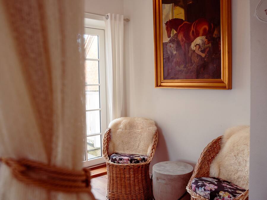 A room with two wicker chairs, one with a floral cushion and another with a fur throw. A painting hangs on the wall, and a window with curtains is visible.