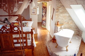 A bedroom with a wooden bed, a white bathtub, and a stone wall. - Boutique Hotel Postgården (Holsted)
