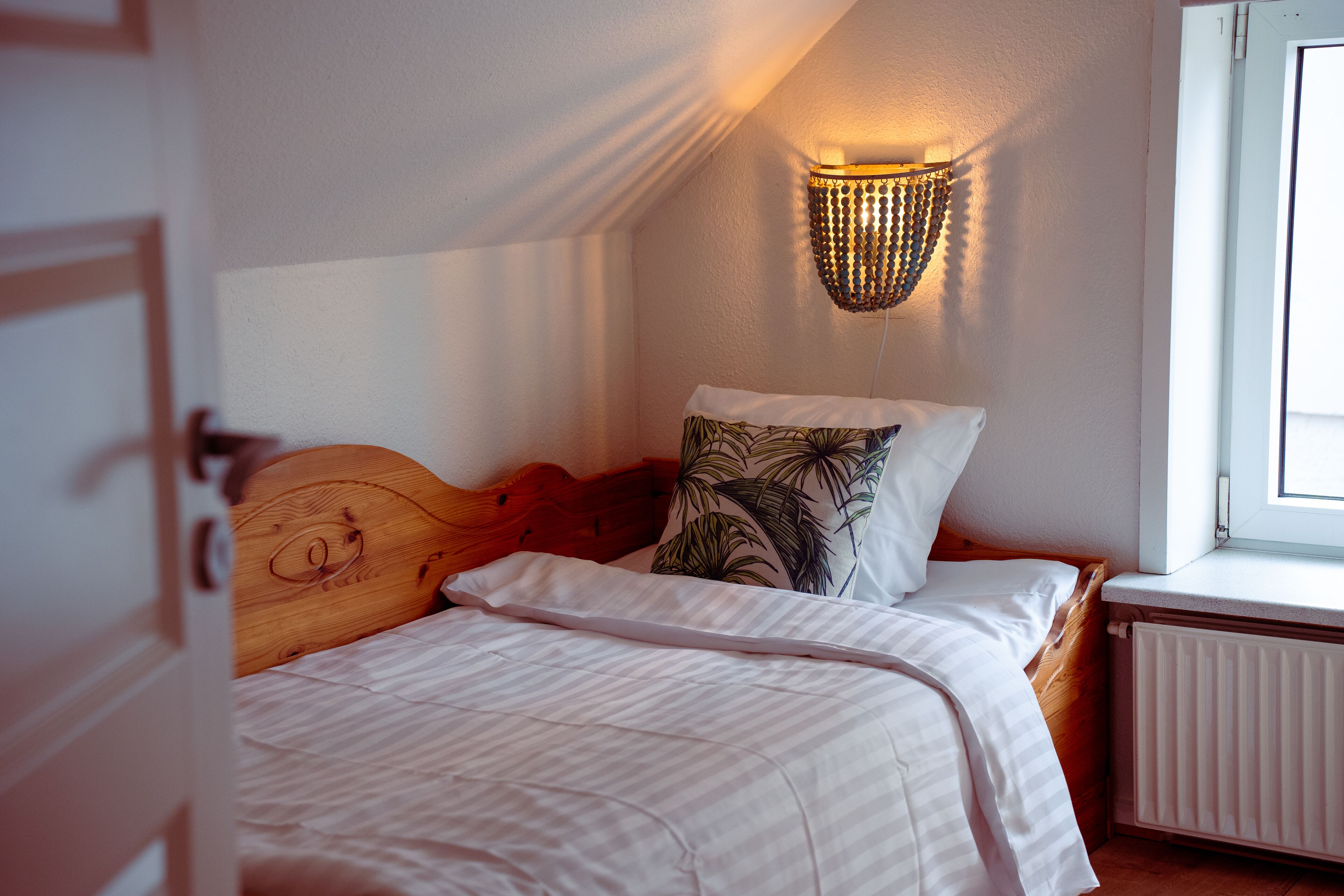 Un dormitorio con cabecera de madera, cama con ropa de cama blanca, almohada estampada, lámpara de pared y ventana con vistas.