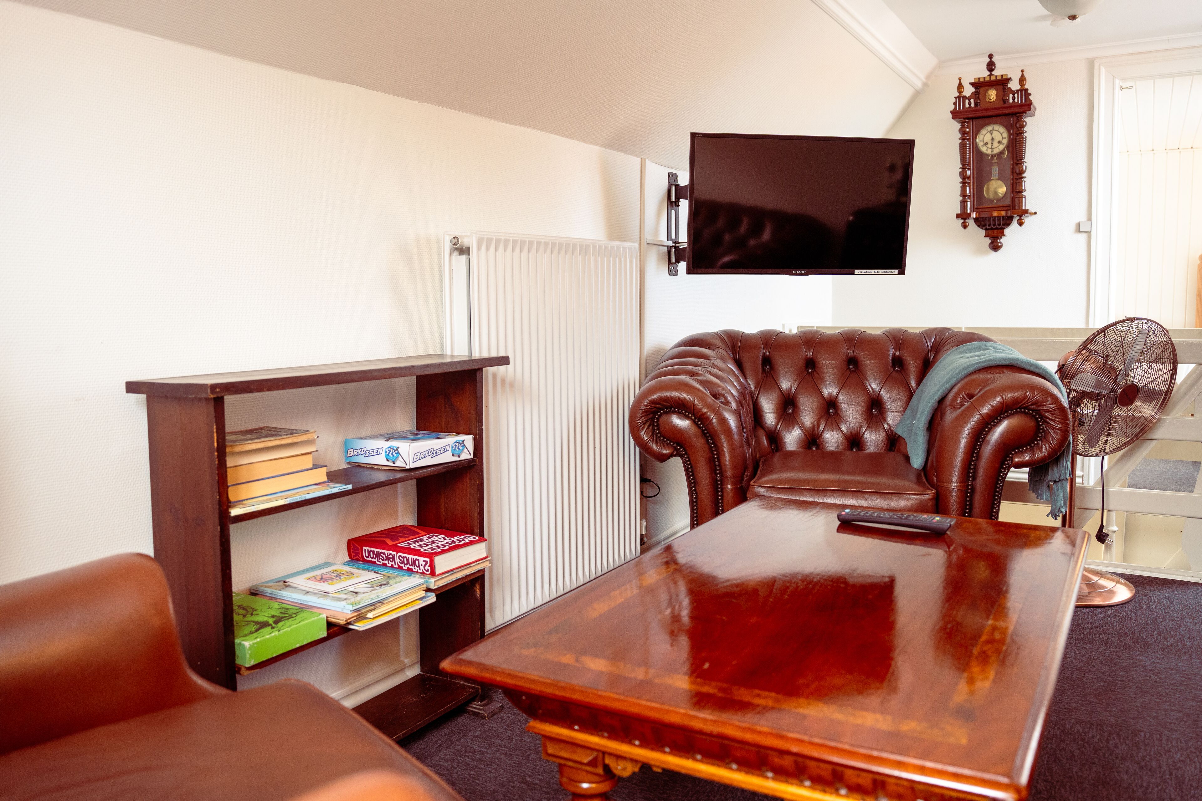 Una sala de estar con un sofá de cuero marrón, una mesa de centro de madera, una estantería con libros, un televisor montado en la pared y un reloj de pared.