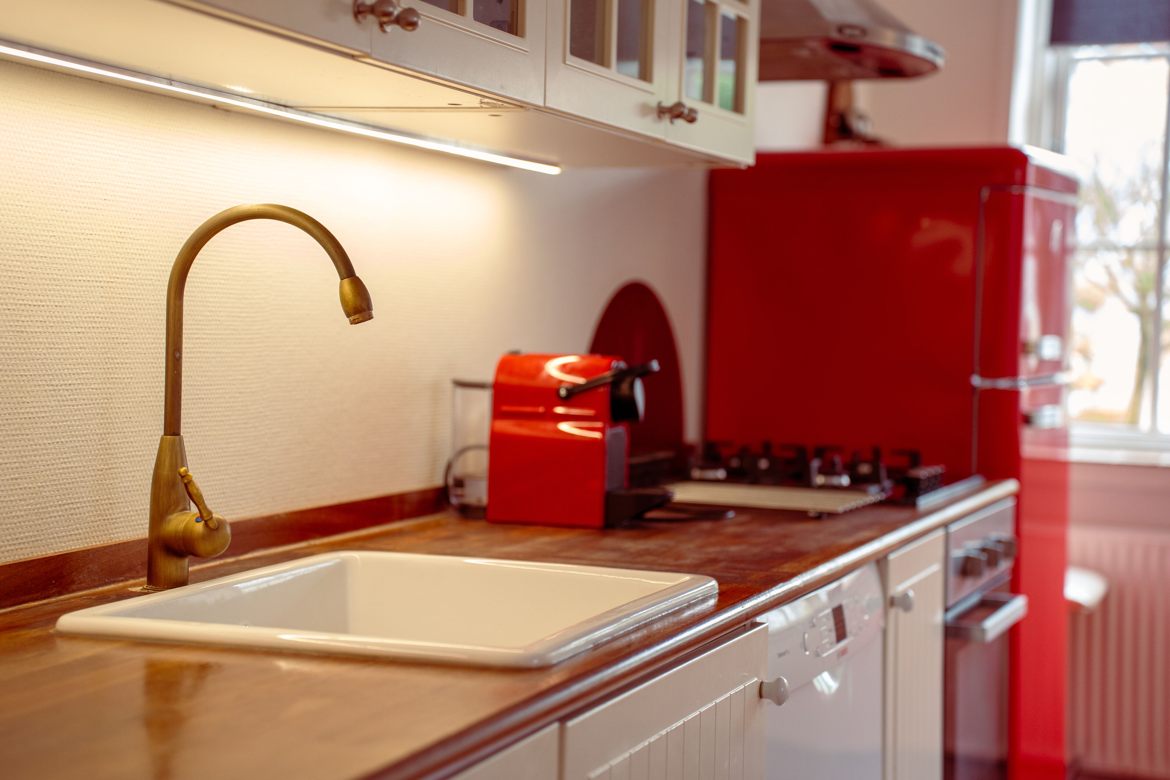 A kitchen with a white sink, a brass faucet, a red appliance, and wooden cabinets.