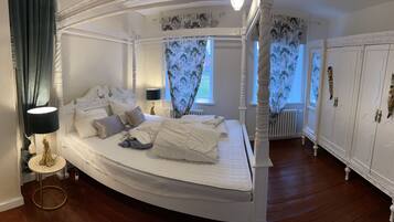 A four-poster bed with a white canopy, a round gold table with a lamp, and a white wardrobe with decorative handles.
