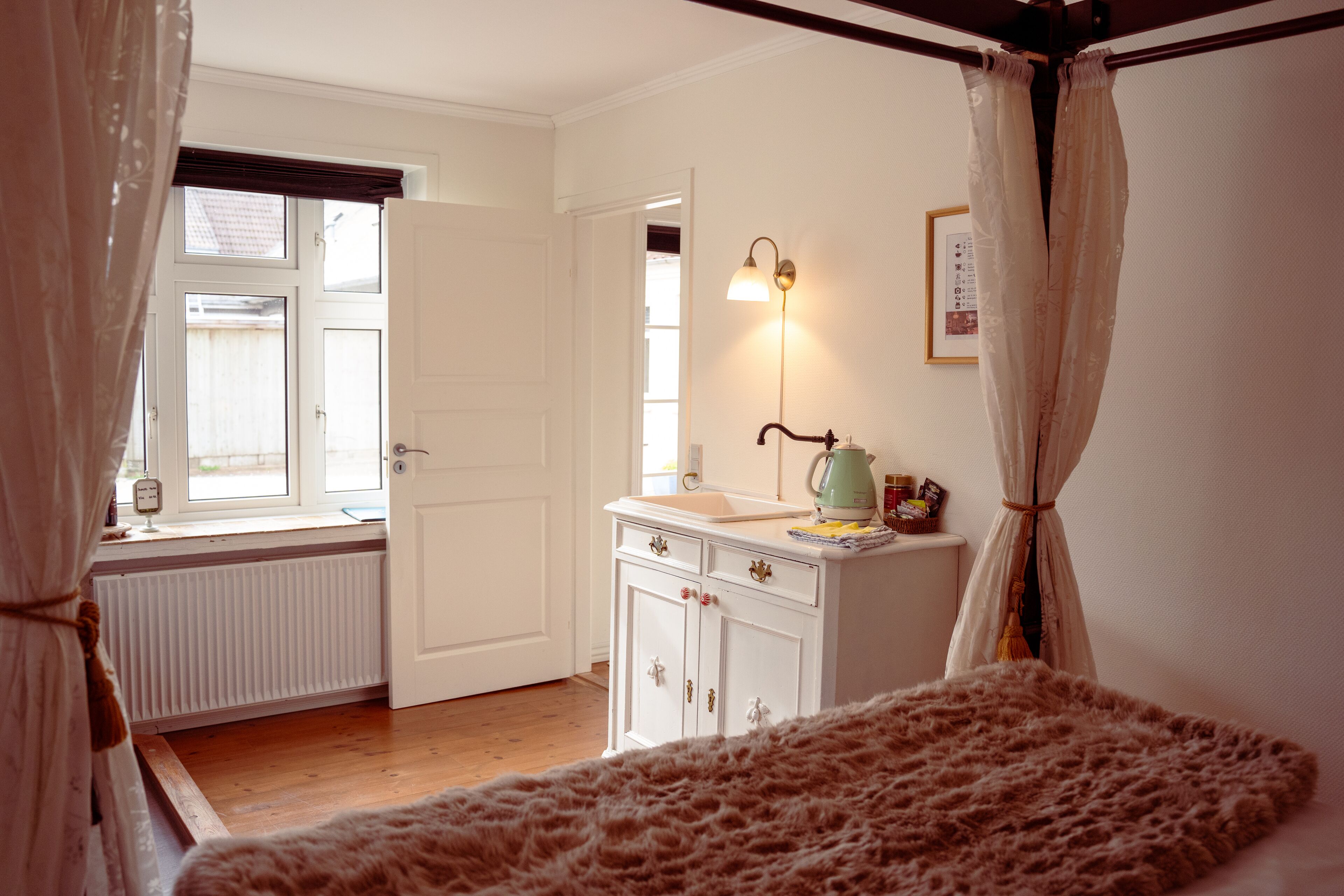 A bedroom with a wooden floor, a white dresser with drawers, a bedside table with a lamp, and a window with curtains.