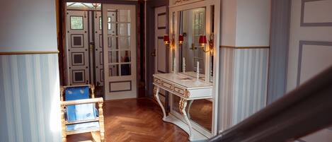 Un pasillo con suelo de madera, una silla azul, una consola blanca con un espejo, y una puerta con paneles de vidrio.