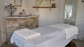 A spa room with a stone fireplace, a bed with a white blanket, shelves with rolled towels, and a framed certificate on the wall.
