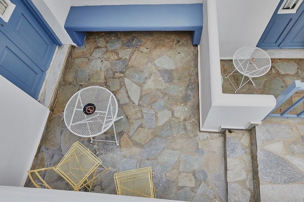 Un patio con suelo de piedra, dos mesas de metal blanco con sillas y un banco azul.