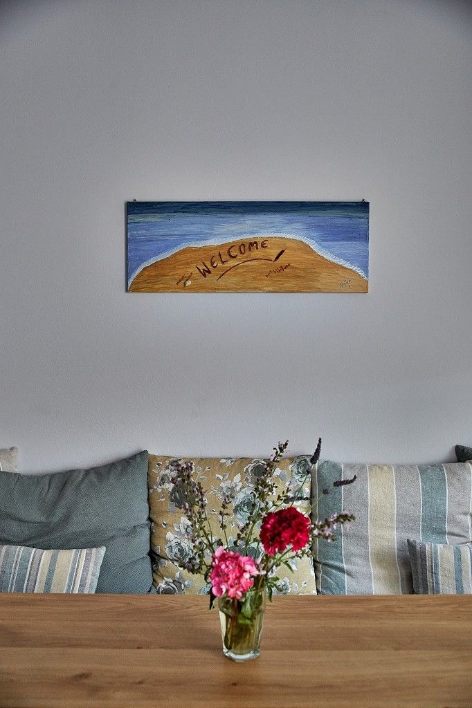 A painting of a beach with the word 'WELCOME' on a wooden board, a vase with flowers on a table, and a sofa with patterned cushions.