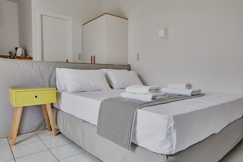 Una cama bien hecha con ropa de cama blanca y una manta de tiras, una mesita de noche pequeña de color amarillo y un espejo sobre la cama.
