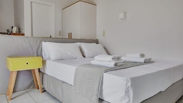 Una cama bien hecha con ropa de cama blanca y una manta de tiras, una mesita de noche pequeña de color amarillo y un espejo sobre la cama.