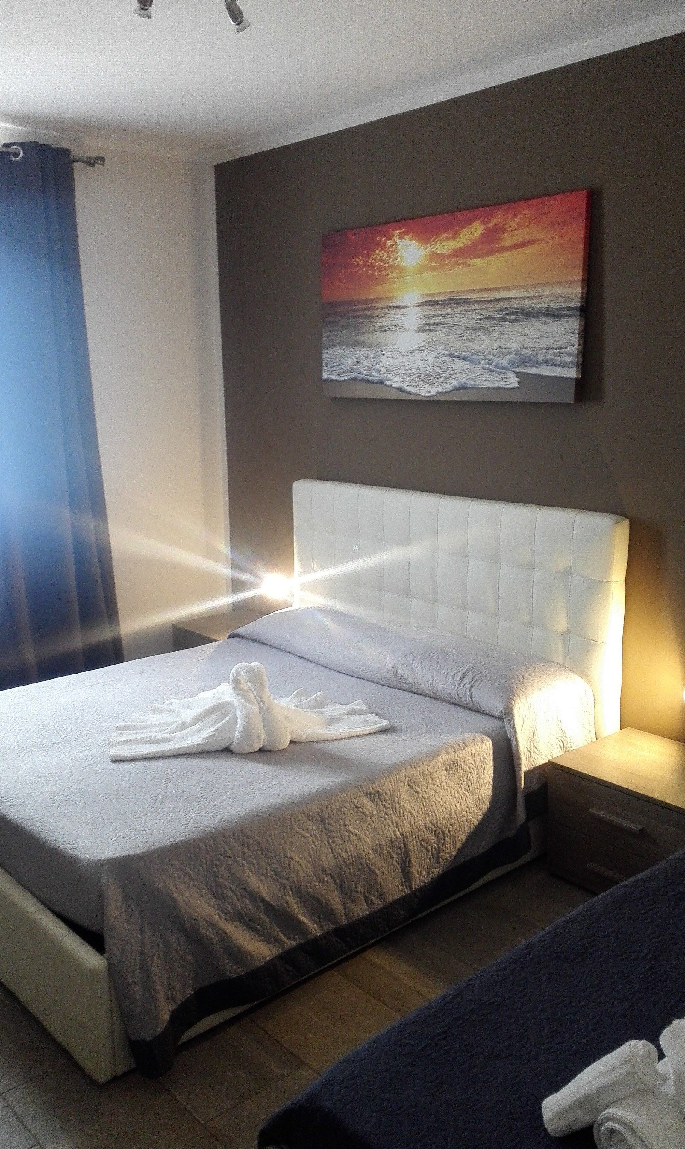 Una habitación de hotel con una cama, un cuadro de una puesta de sol sobre el océano y una cortina azul.