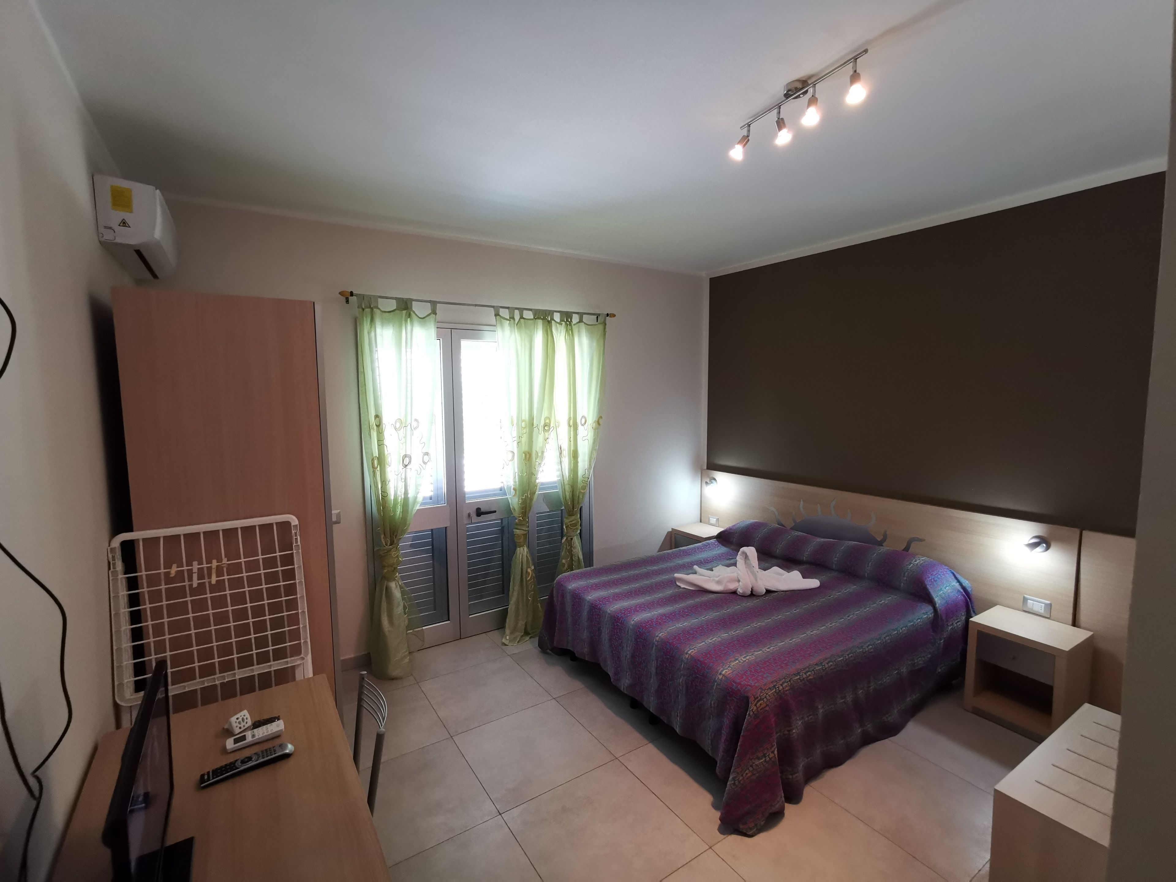 A bedroom with a bed, bedside tables, a desk with a chair, a TV, and a window with curtains.