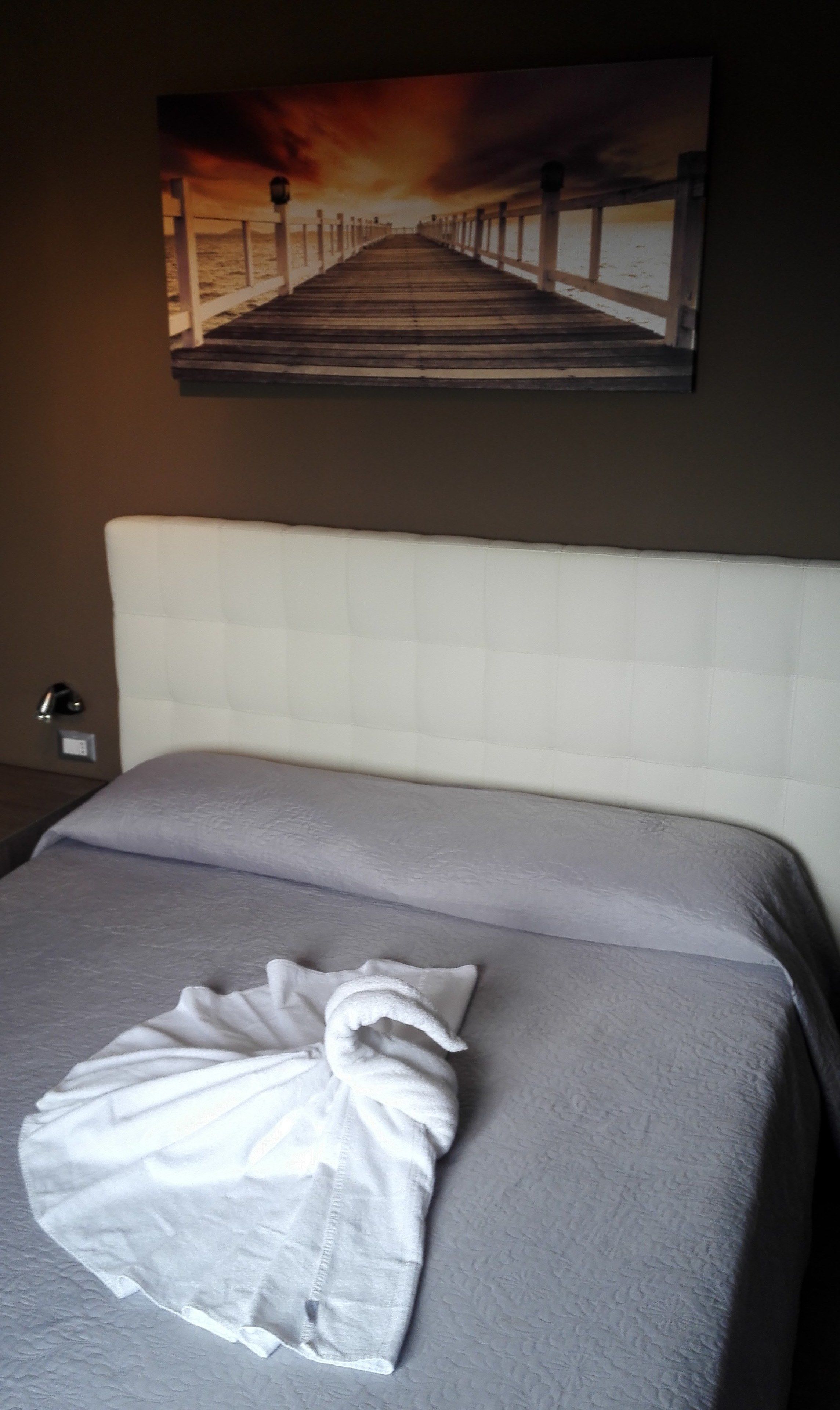 Una habitación de hotel con cama, cabecera blanca y un cuadro de un muelle en la pared.