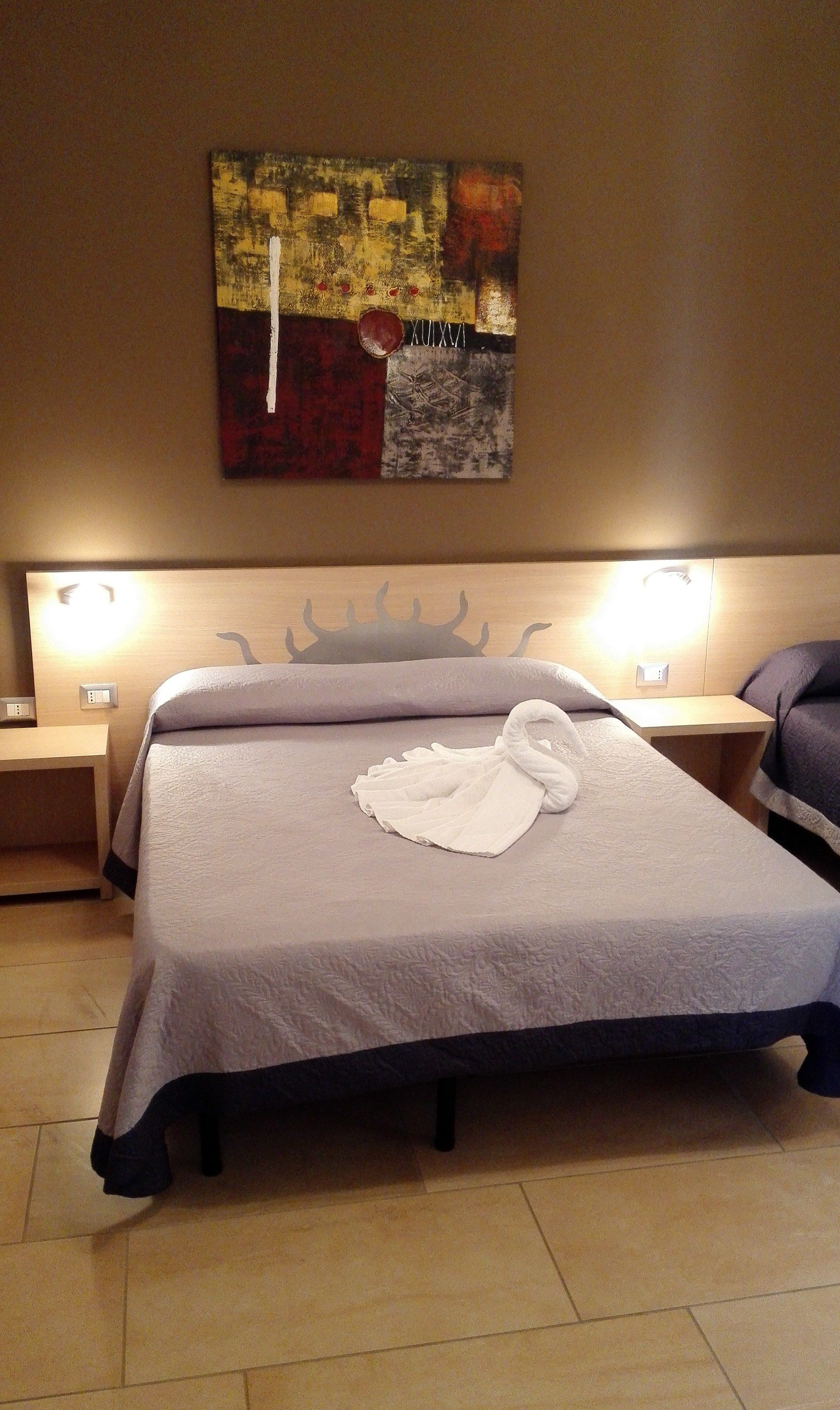 Een hotelkamer met een groot bed, twee nachtkastjes, een aan de muur hangend kunstwerk, en een hoofdbord met een kroonmotief.