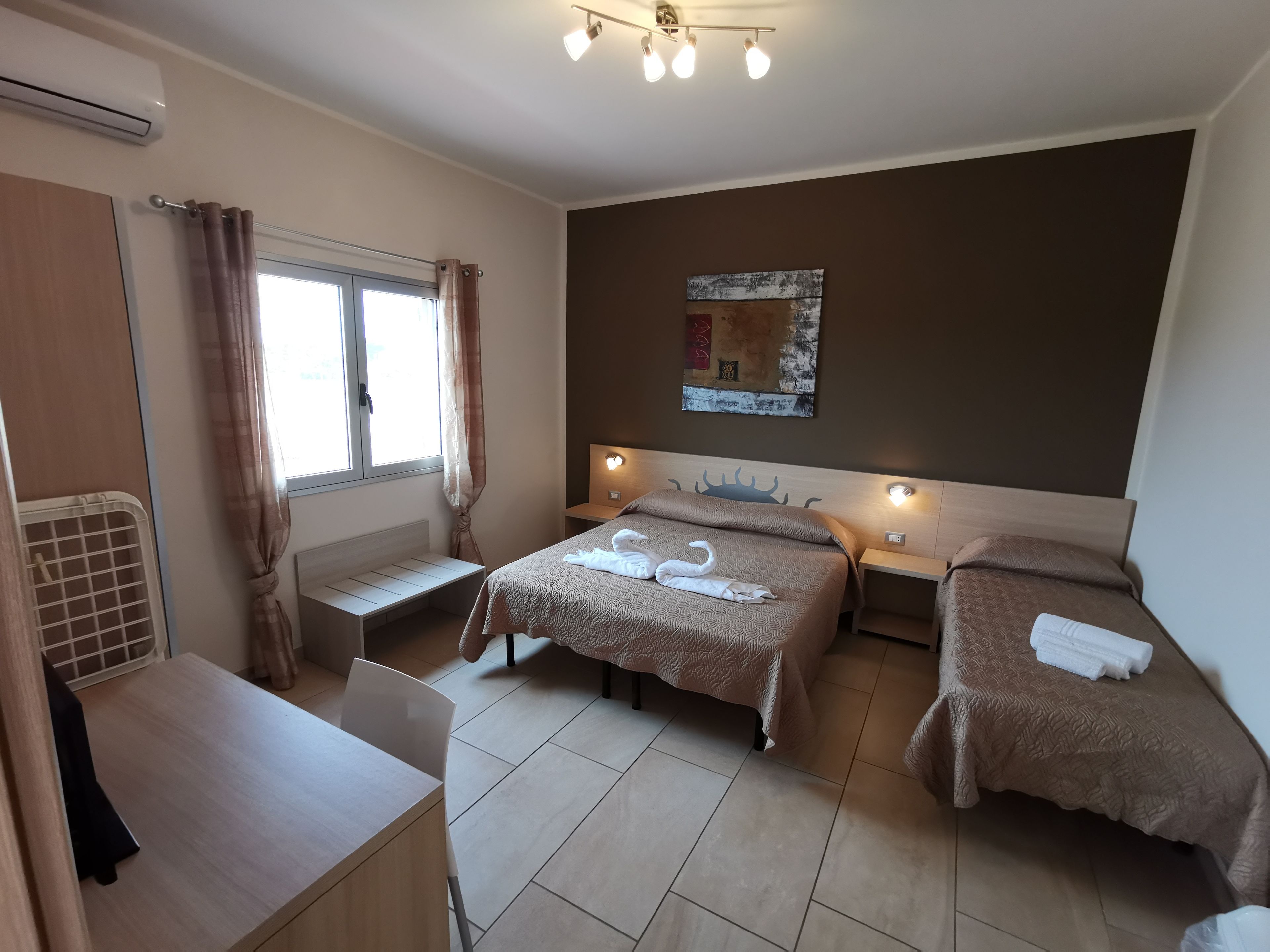 A hotel room with two beds, a desk, a chair, a window with curtains, a wall-mounted light, and a ceiling light fixture.