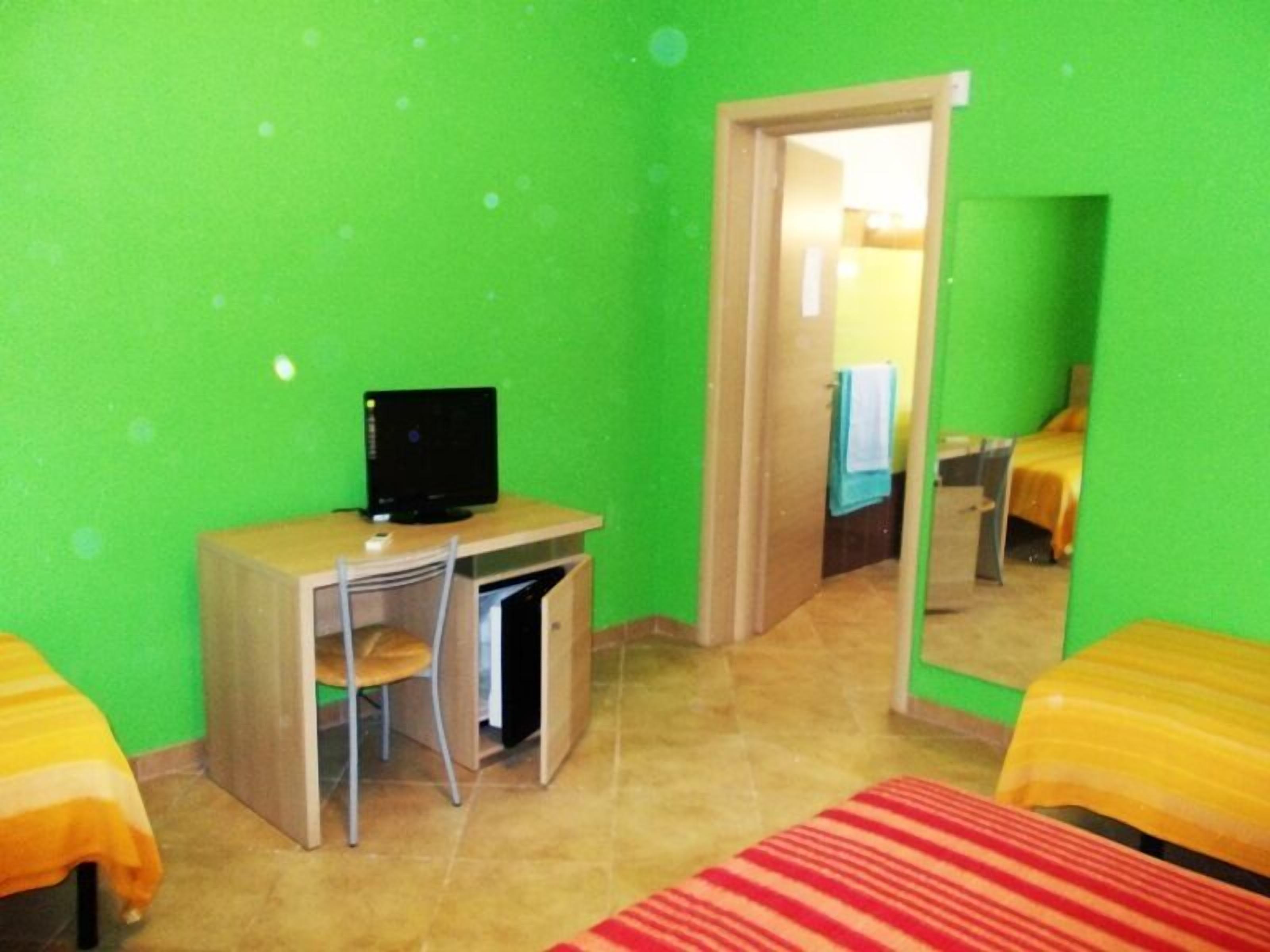 Ein Zimmer mit Schreibtisch, Stuhl und Bett. An der Wand befindet sich ein Spiegel und eine Tür zu einem weiteren Raum.