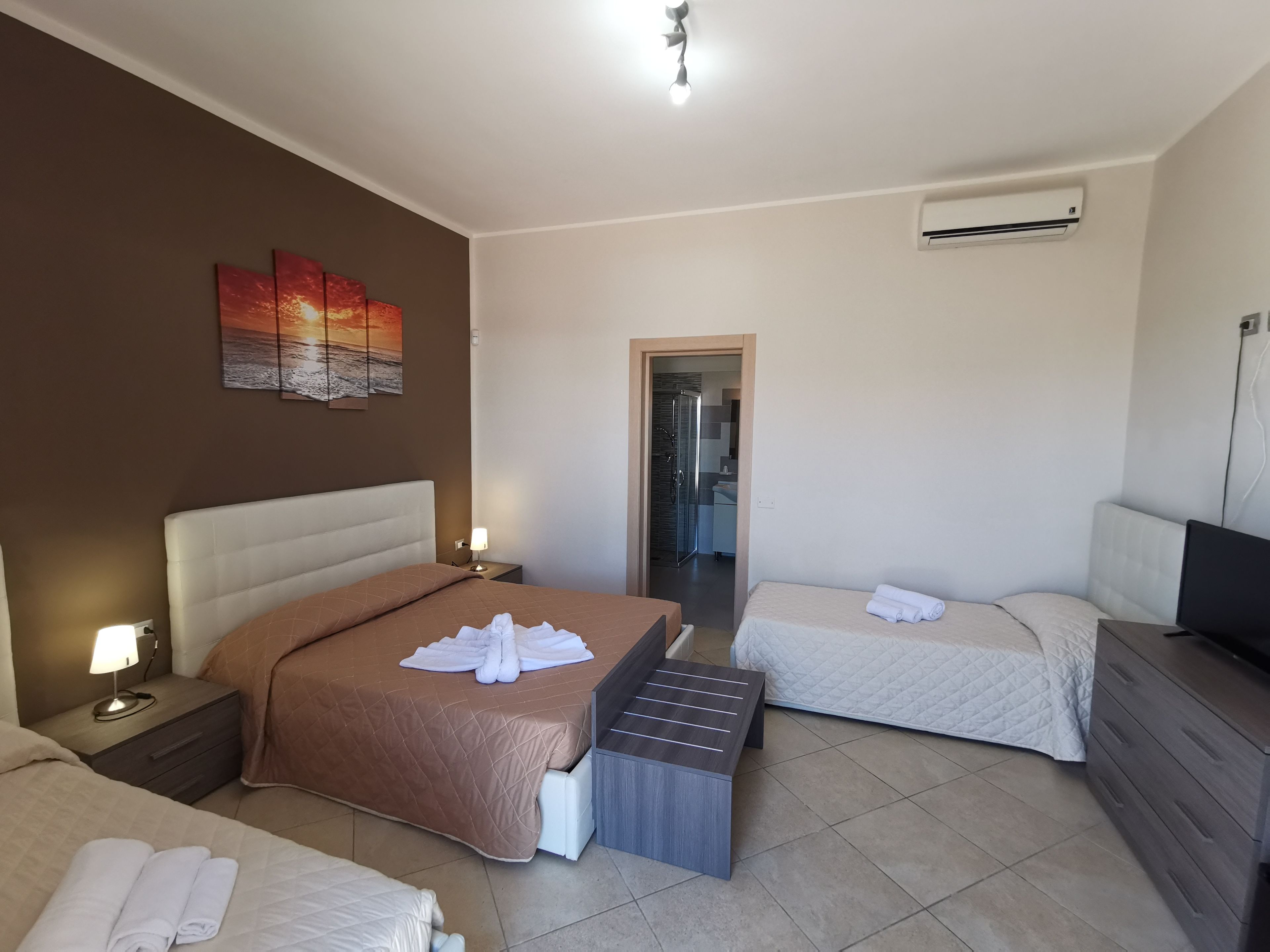 Ein Hotelzimmer mit einem großen Bett, einem Einzelbett, einem Schreibtisch, einem Stuhl, einem Kleiderschrank und einer Klimaanlage. Ein Badezimmer ist durch eine offene Tür sichtbar.