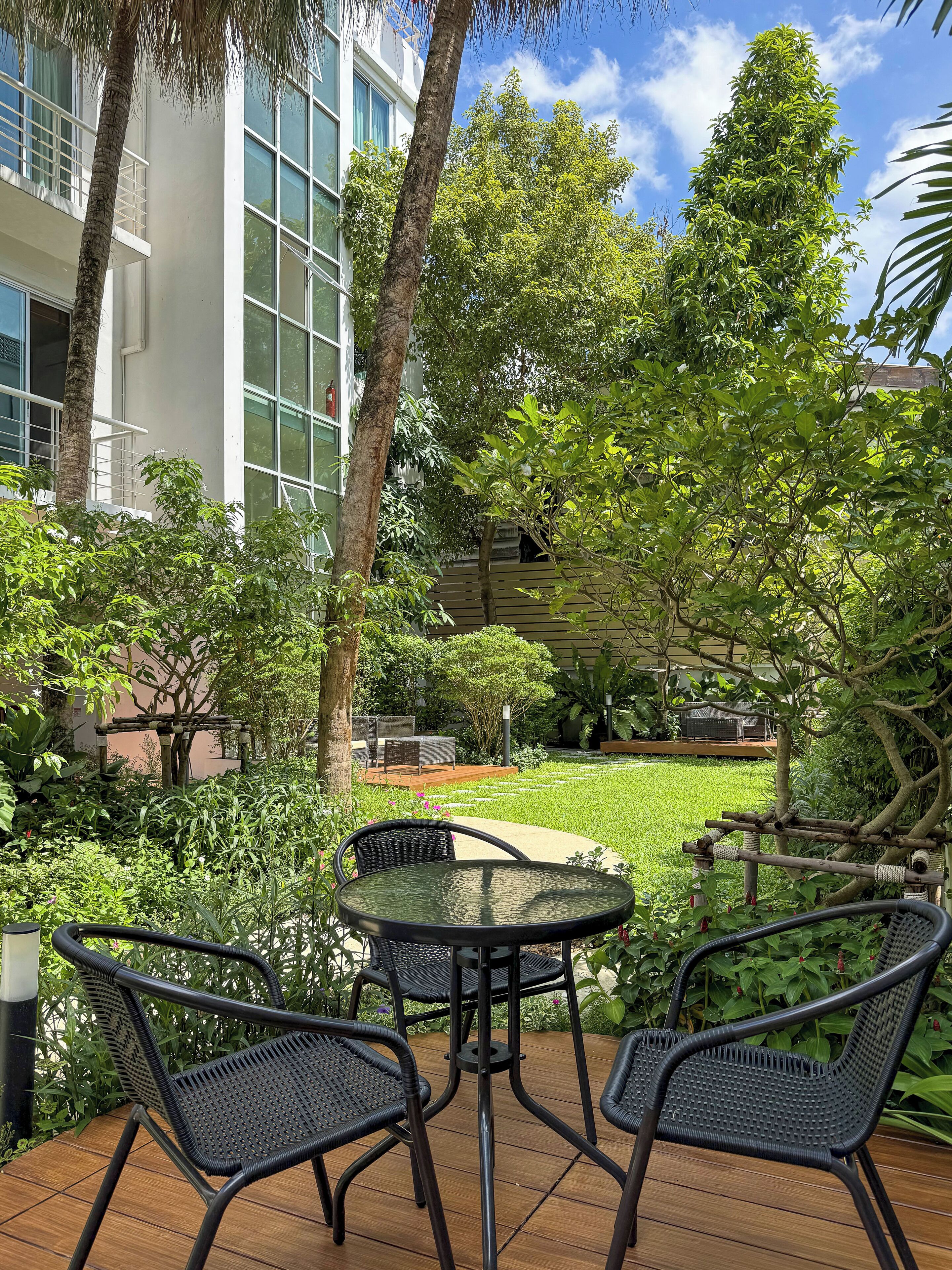 An outdoor patio with a round table and four chairs, surrounded by lush greenery and a building with large windows.