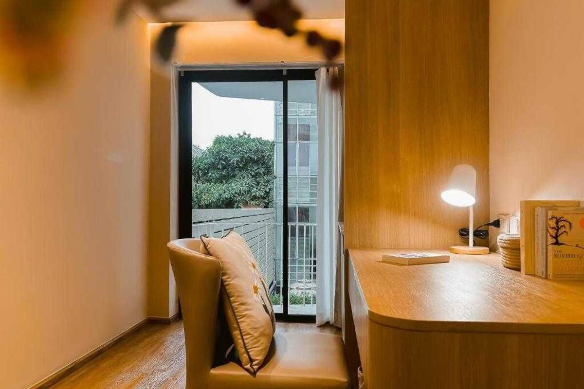 Hotellihuone, jossa on puinen työpöytä, tuoli, pieni pöytä lampun kanssa ja näkymä lasiovesta ulkoalueelle, jossa on viherkasveja.