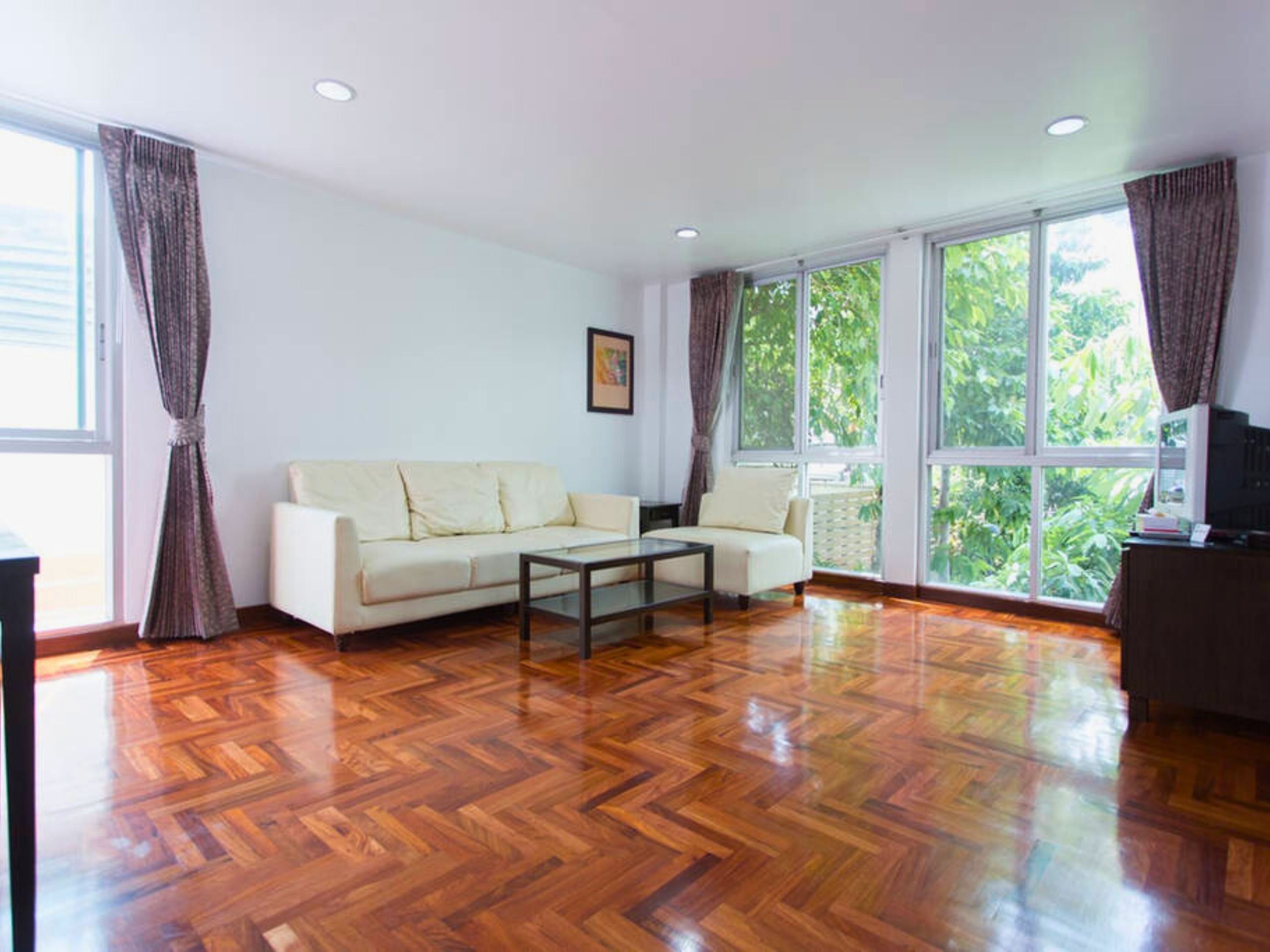 A spacious living room with a wooden parquet floor, a white sofa, a glass coffee table, and large windows offering a view of greenery outside.