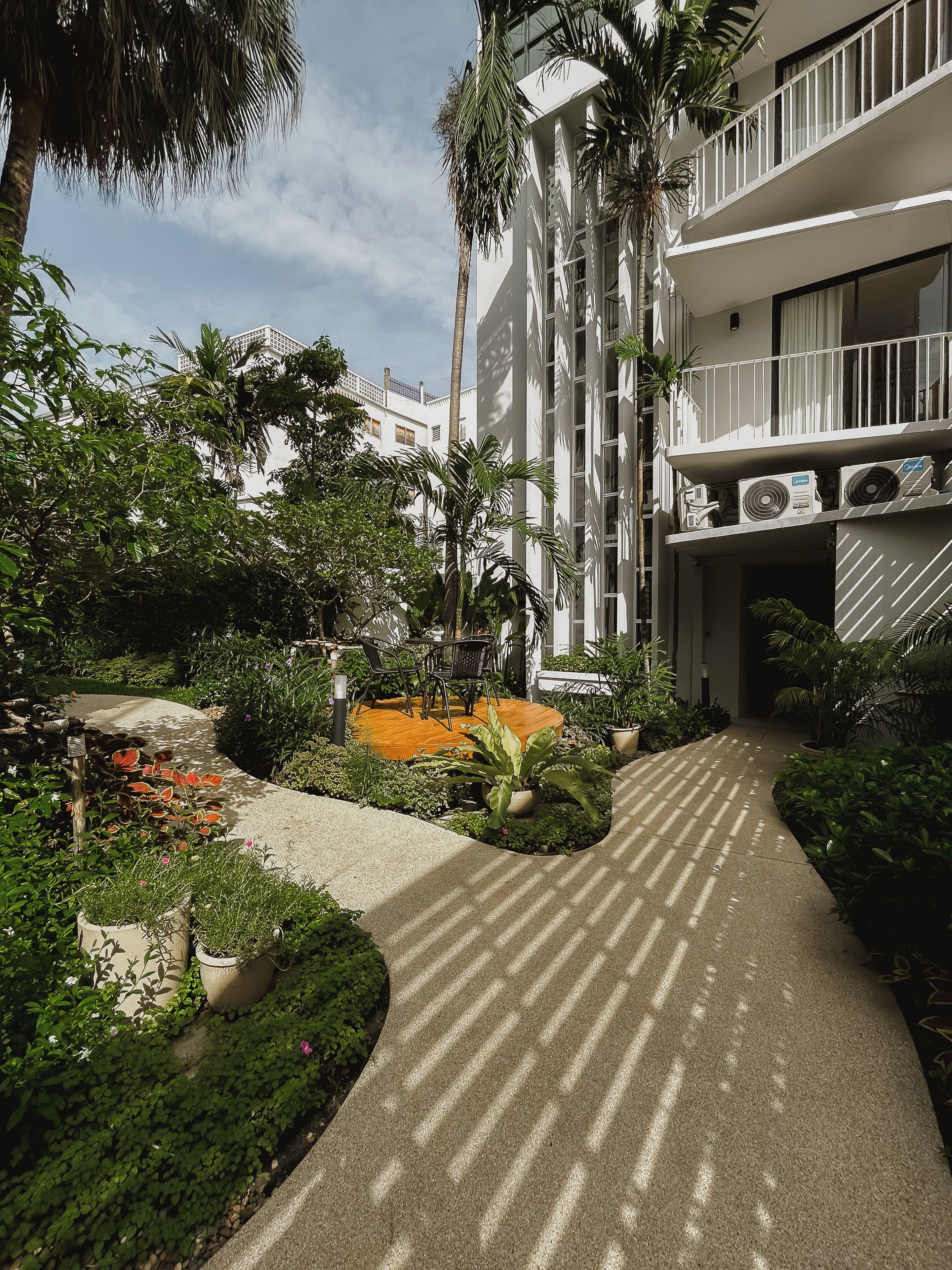 A pathway leading to a building with multiple air conditioning units on the exterior, surrounded by lush greenery and potted plants.